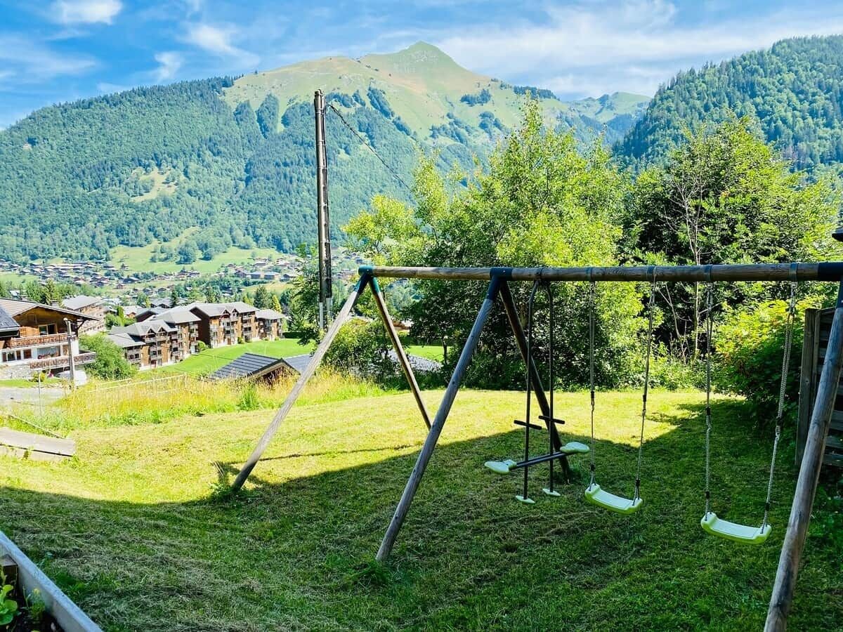 Un espacio verde con un columpio, una cerca de madera y vistas a una montaña y edificios al fondo.