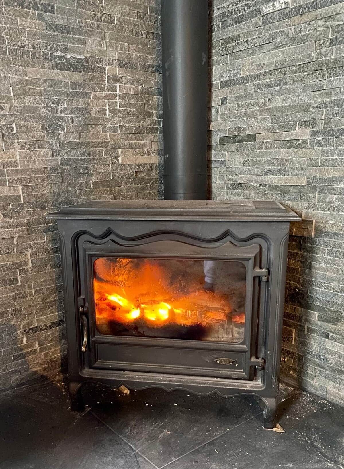 A wood-burning stove with a glass door, set against a brick wall with a black pipe above it.