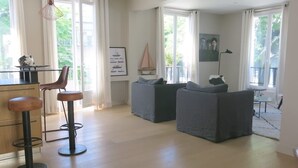 A spacious living room with a large window, a dining area with a table and chairs, and a kitchen area with a bar stool. - Sacré Coeur CityCosy (Paris)