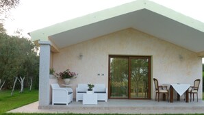 Une terrasse couverte, meublée de mobilier blanc, avec une table et des chaises, entourée d’un jardin.