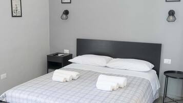 Un letto rifatto con lenzuola bianche e asciugamani arrotolati, un comodino con telecomando e un portalampada fissato alla parete.