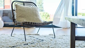 A wicker chair with a fluffy pillow, a white shag rug, and a view of greenery through sheer curtains.
