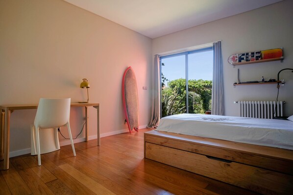 A bedroom with a wooden bed, a desk with a chair, a surfboard, and a view of greenery through a large window.