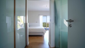 A modern bedroom with a wooden floor, a bed with white bedding, and a view of the outdoors through a large window.