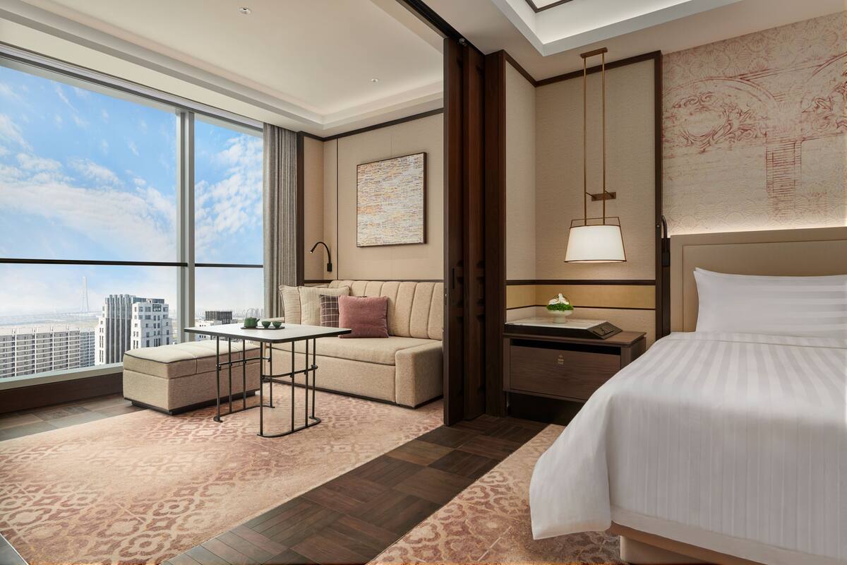 A modern hotel room with a large window, a sofa, a desk, and a bed.