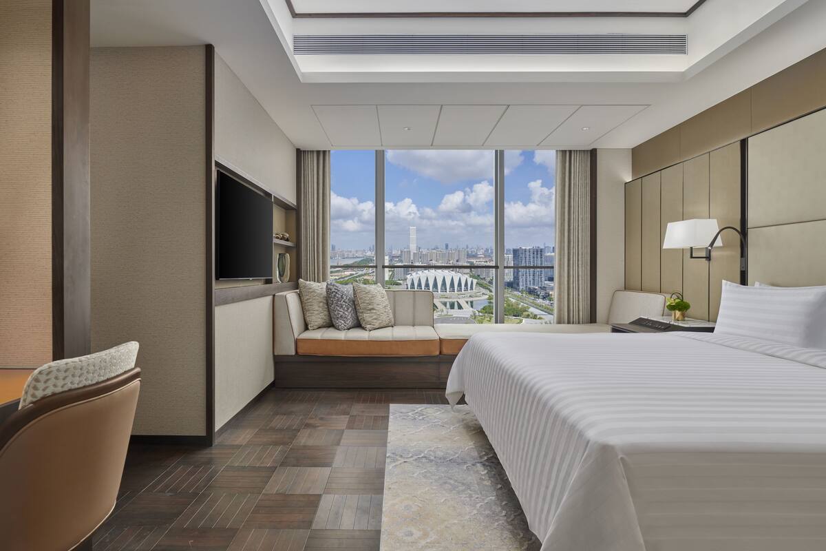 A modern hotel room with a large bed, a seating area with a TV, and a city view.