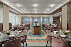 A hotel dining area with multiple tables and chairs, a central decorative column, and a patterned carpet.