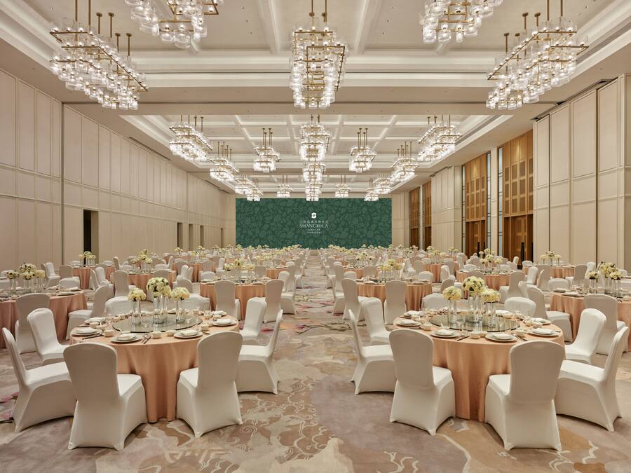 Um salão de banquetes com mesas redondas, cadeiras brancas e lustres.