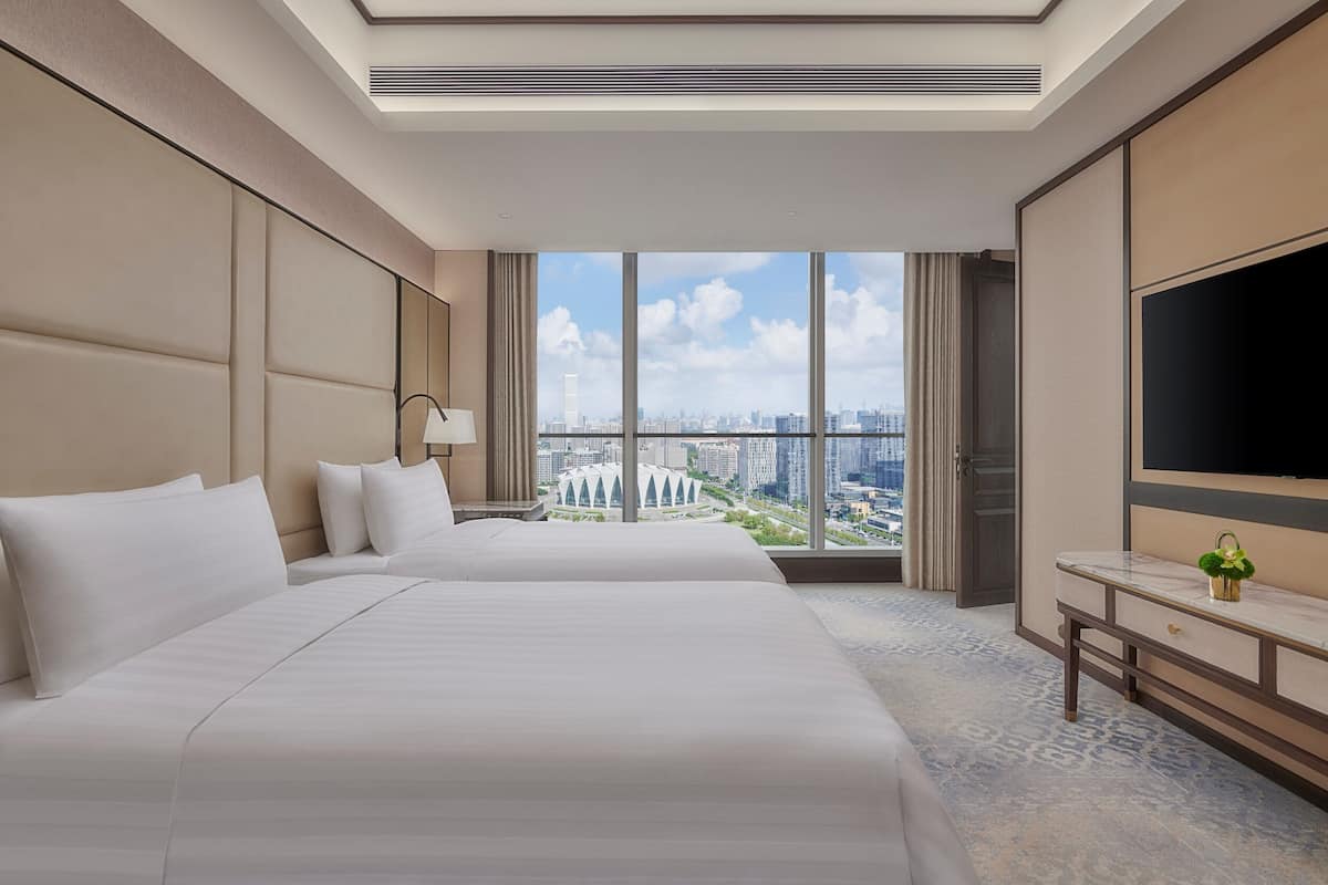 A hotel room with two beds, a large window with a city view, a flat-screen TV, and a desk with a lamp.
