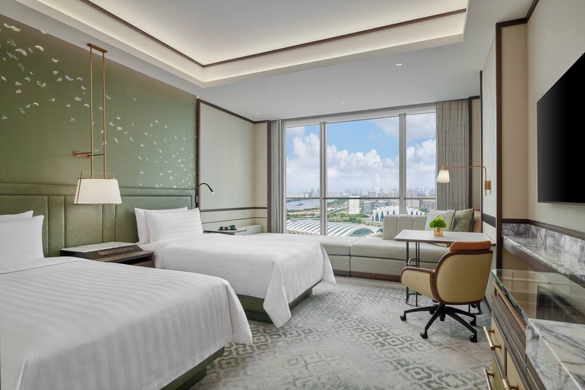 A hotel room with two beds, a desk, a chair, a TV, and a large window with a city view.