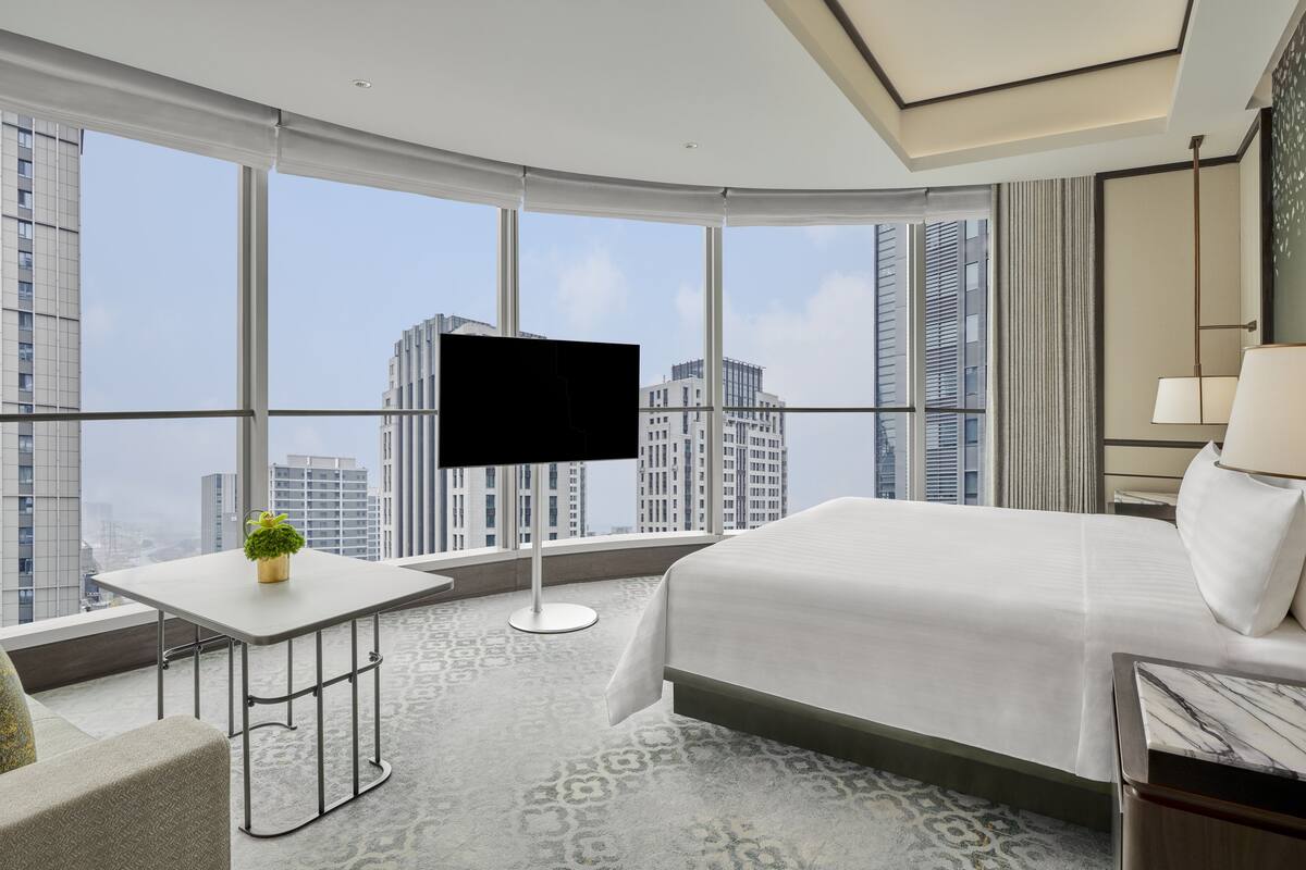A modern hotel room with a large bed, a flat-screen TV, a small table with a plant, and a view of city buildings through large windows.
