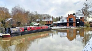 A narrow canal with a narrowboat named "SMOKE" docked on the left. There are buildings on the right bank, and the water reflects the buildings and the sky.