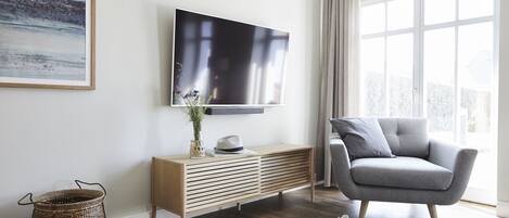 En moderne stue med et fladskærms-tv på væggen, en grå sofa, et træbord med en vase og en hat, en fletkurv og et stort vindue med gardiner.