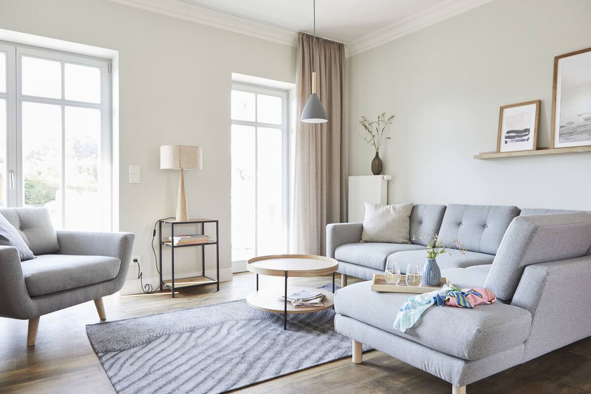 A modern living room with a grey sofa, a round coffee table, and a grey armchair. There is a window with curtains, a floor lamp, and a vase with flowers on a shelf.