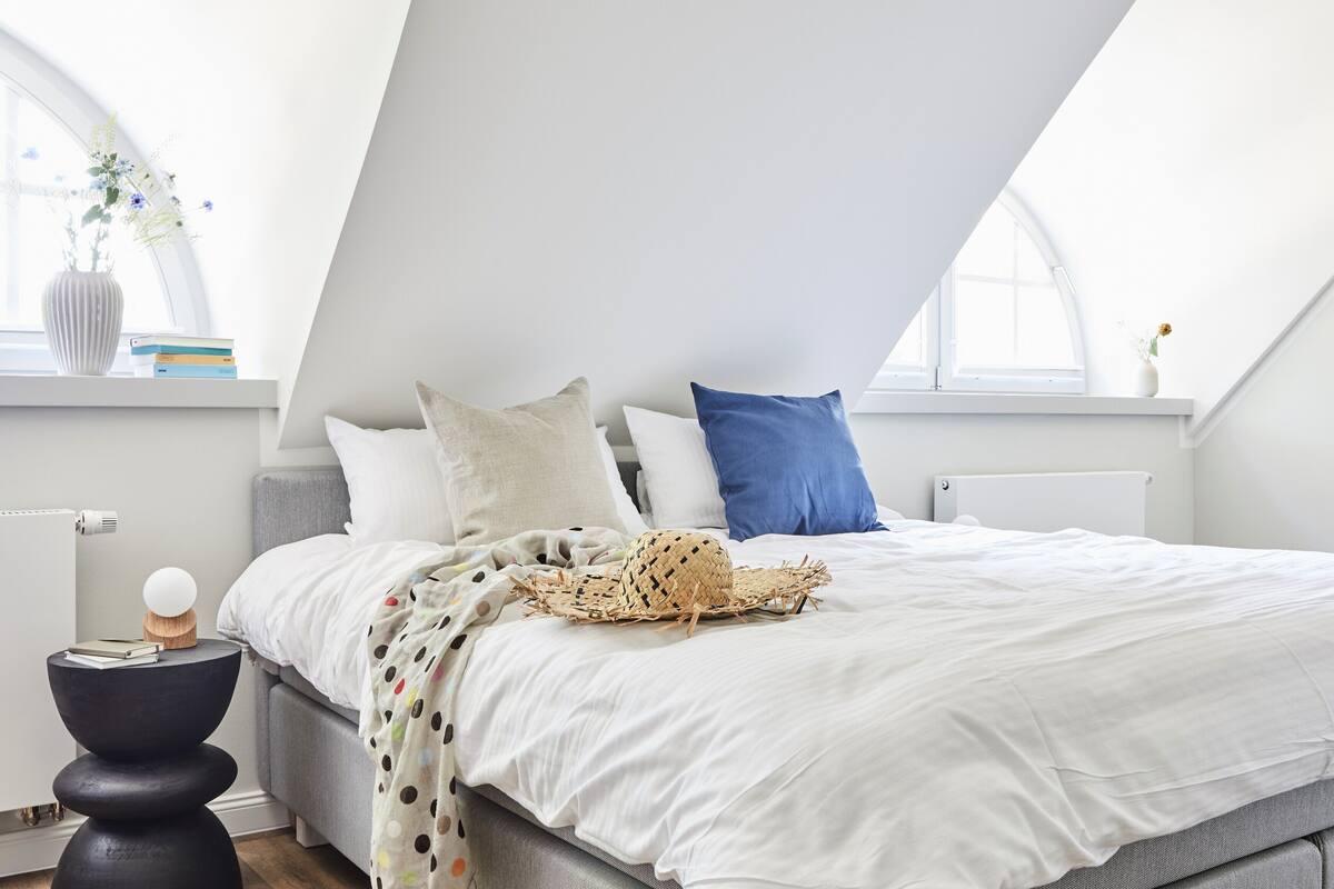 A modern bedroom with a sloped ceiling, a bed with white bedding, a blue pillow, and a woven basket on the bed.