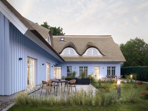 Una casa azul con techo de paja, un patio con sillas y un jardín con plantas y flores.