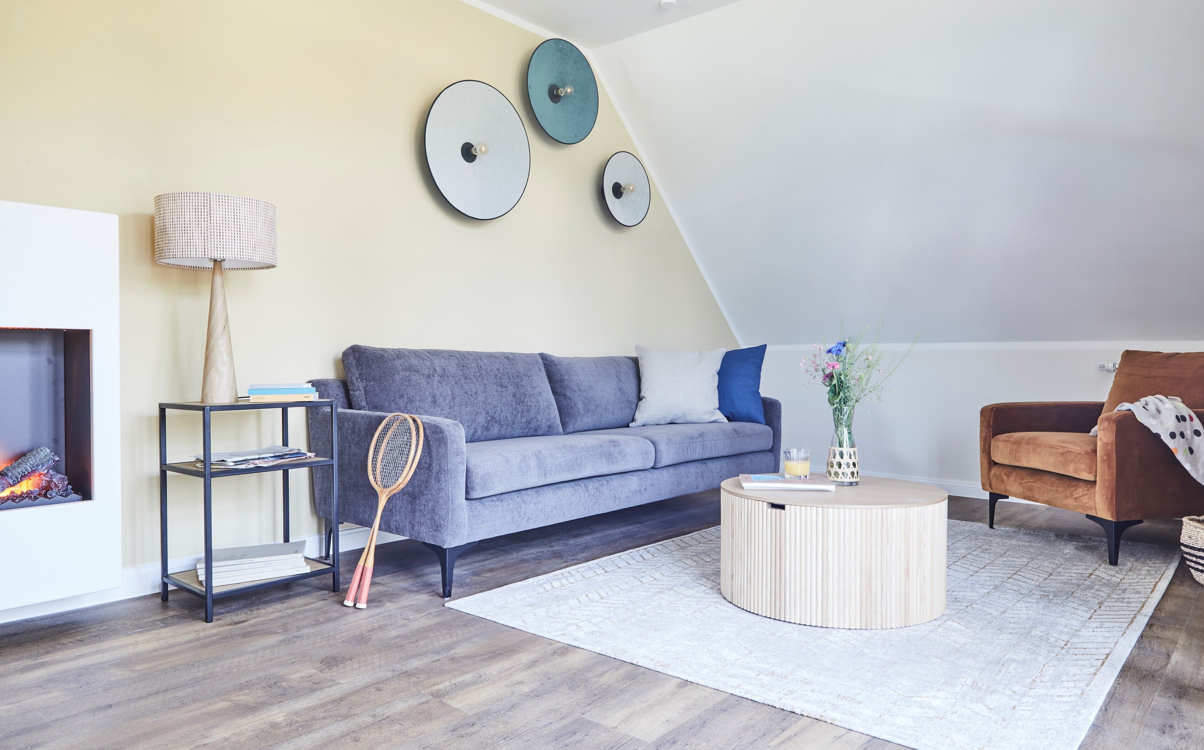 A modern living room with a grey sofa, a round coffee table, a fireplace, and a wall-mounted clock.