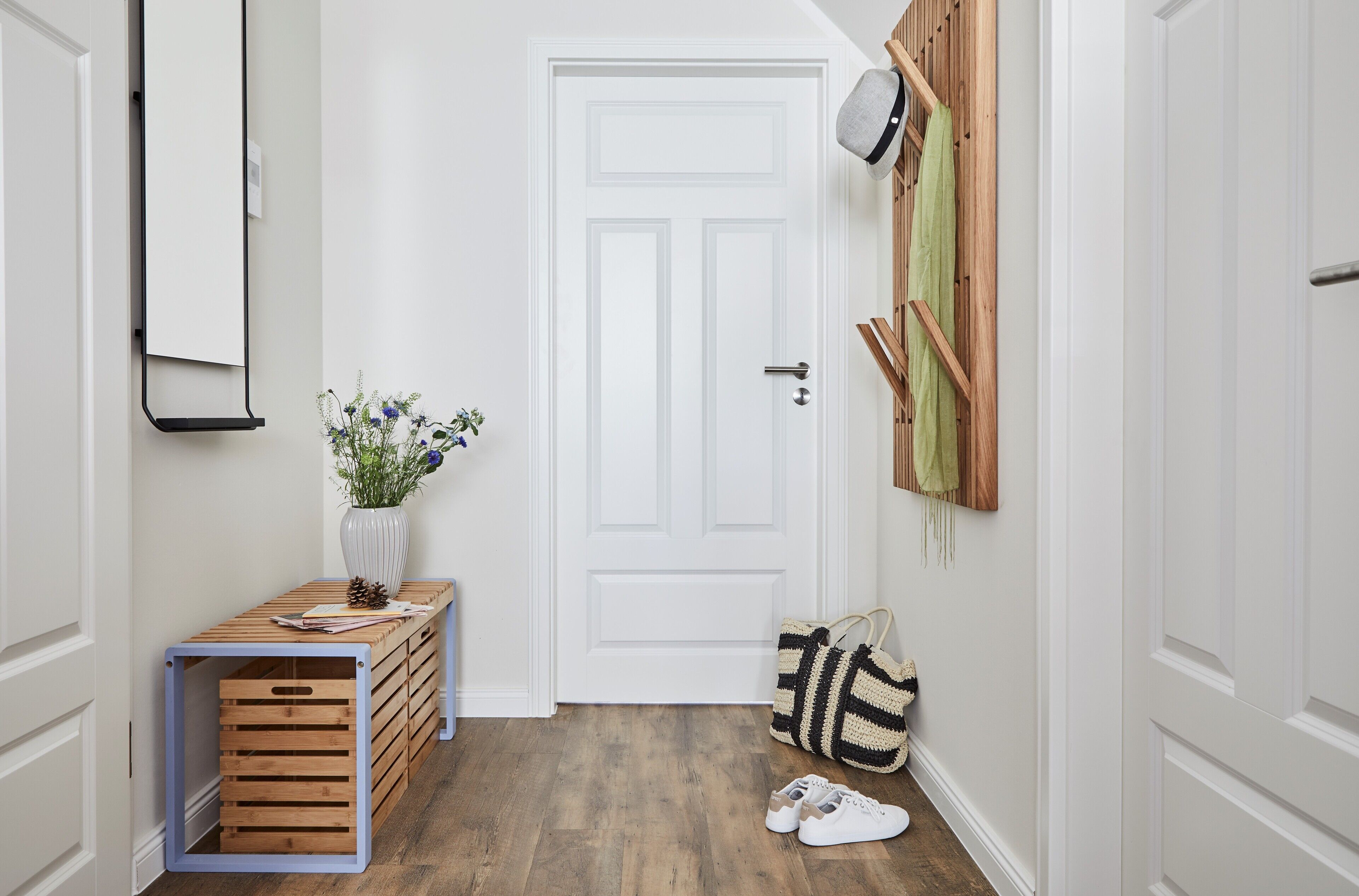 Un couloir avec une porte blanche, un banc en bois sur lequel est posé un vase de fleurs, et un sac à rayures sur le sol.