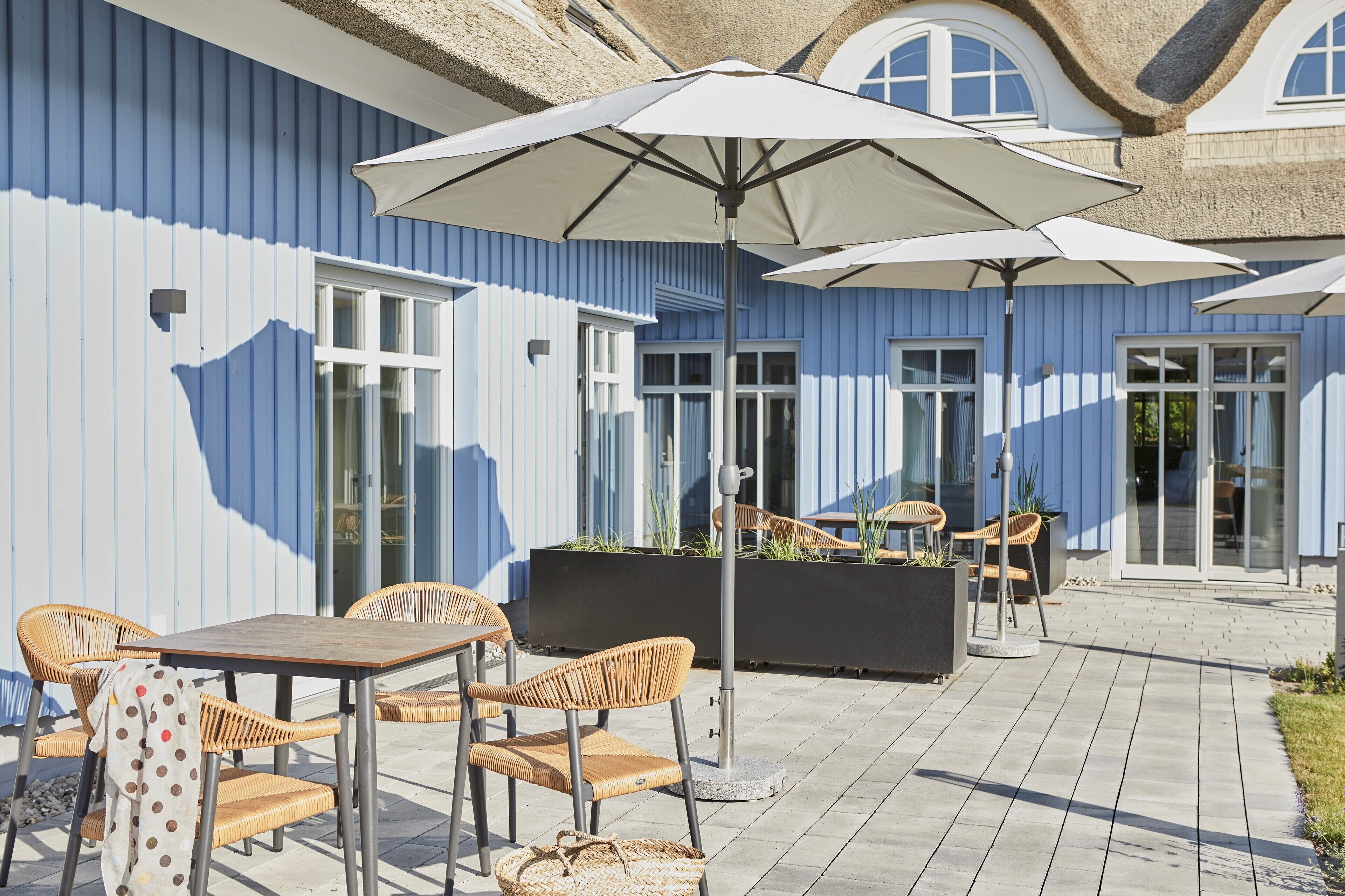 An outdoor patio with wicker chairs and tables, umbrellas, and a planter box with plants.
