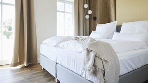Una cama bien hecha con sábanas blancas y una manta gris, un cabecero de listones de madera y un ventanal con cortinas.