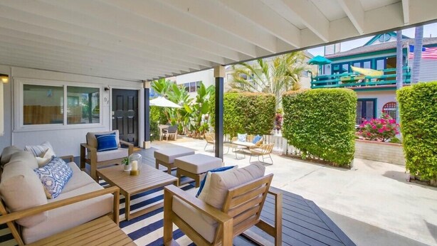 An outdoor patio with wooden furniture, a striped rug, and a view of a building with a balcony. - Convenient Ground Level * Steps to Ocean * Outdoor Patio (San Diego)
