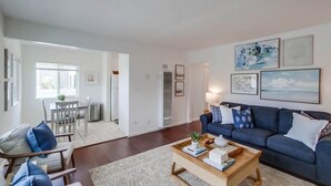 A modern living room with a dark blue sofa, a wooden coffee table, and a dining area with a white table and chairs. - Steps to Ocean + Bright Top Level Unit + Sun Deck (San Diego)