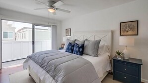 A bedroom with a bed, a ceiling fan, a nightstand, a lamp, and a window with a view of buildings. - Steps to Ocean + Bright Top Level Unit + Sun Deck (San Diego)
