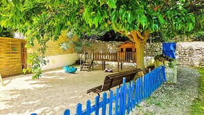 Uma área externa rústica com um banco de madeira, um pequeno casebre e um muro de pedra.