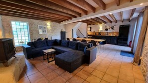 A spacious living area with a dark blue sectional sofa, a small wooden table, and a black fireplace. A kitchen with modern appliances and a staircase leading to an upper level is visible in the background. - The Plough House with 4 bedrooms (Saint-Just-Luzac)