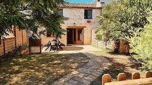 Un edificio de piedra con techo de tejas, una valla de madera y un patio empedrado con mobiliario exterior.