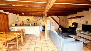 Un interior rústico con una mesa de comedor de madera, un sofá, un televisor y una zona de cocina con pared de piedra y vigas de madera.