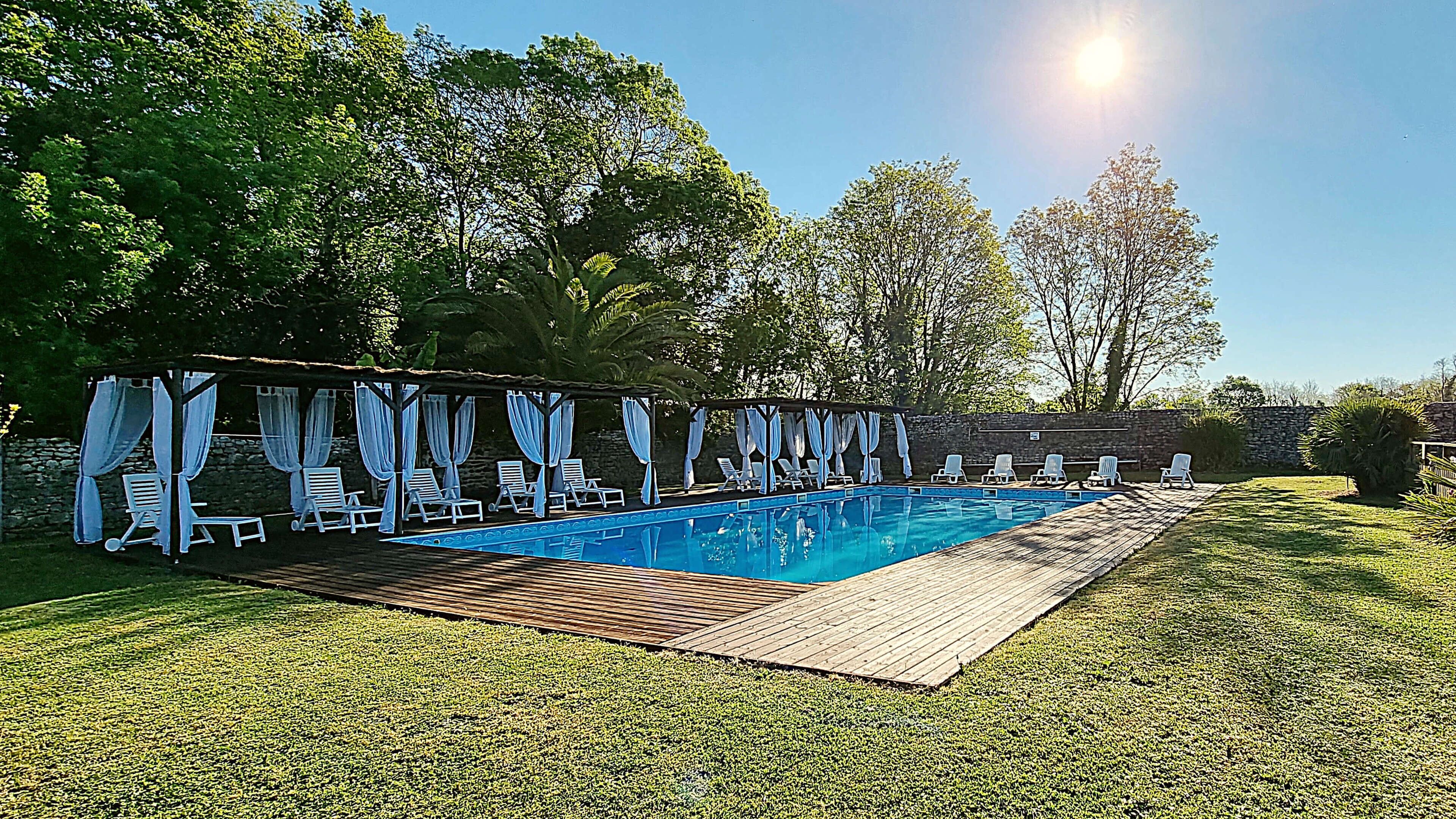 A swimming pool with a wooden deck, surrounded by white lounge chairs and blue curtains.