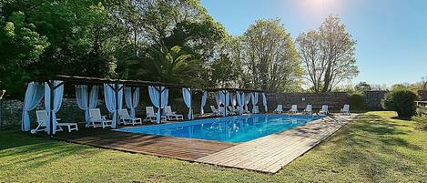 A swimming pool with a wooden deck, surrounded by white lounge chairs and blue curtains.