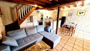 A cozy living room with a grey sectional sofa, a wooden staircase, a kitchen area, and a dining table with chairs. - La Mangeoire 2 bedroom gîte (Saint-Just-Luzac)