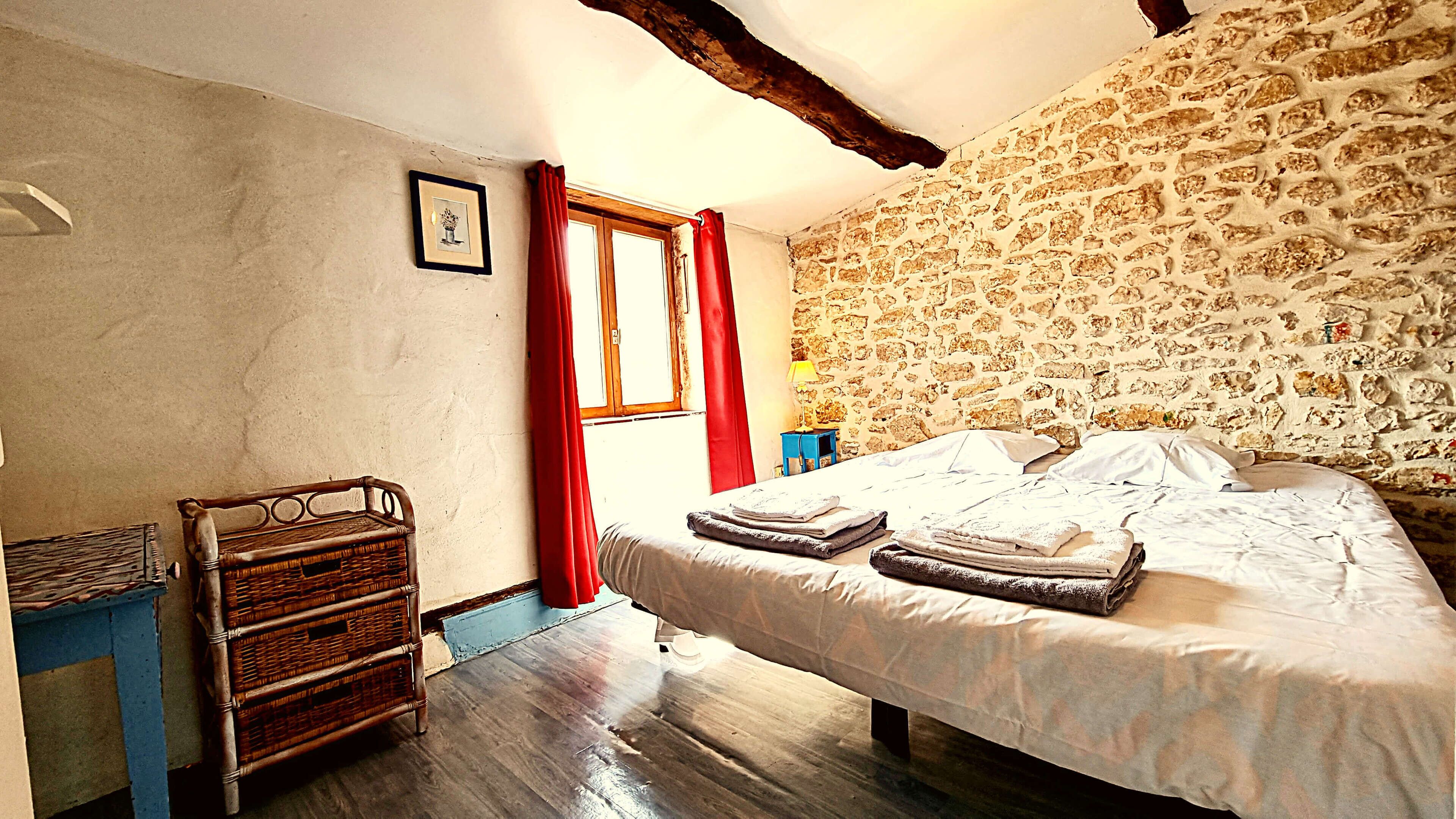 A bedroom with a stone wall, a wooden bed, a wicker chair, and a window with red curtains.
