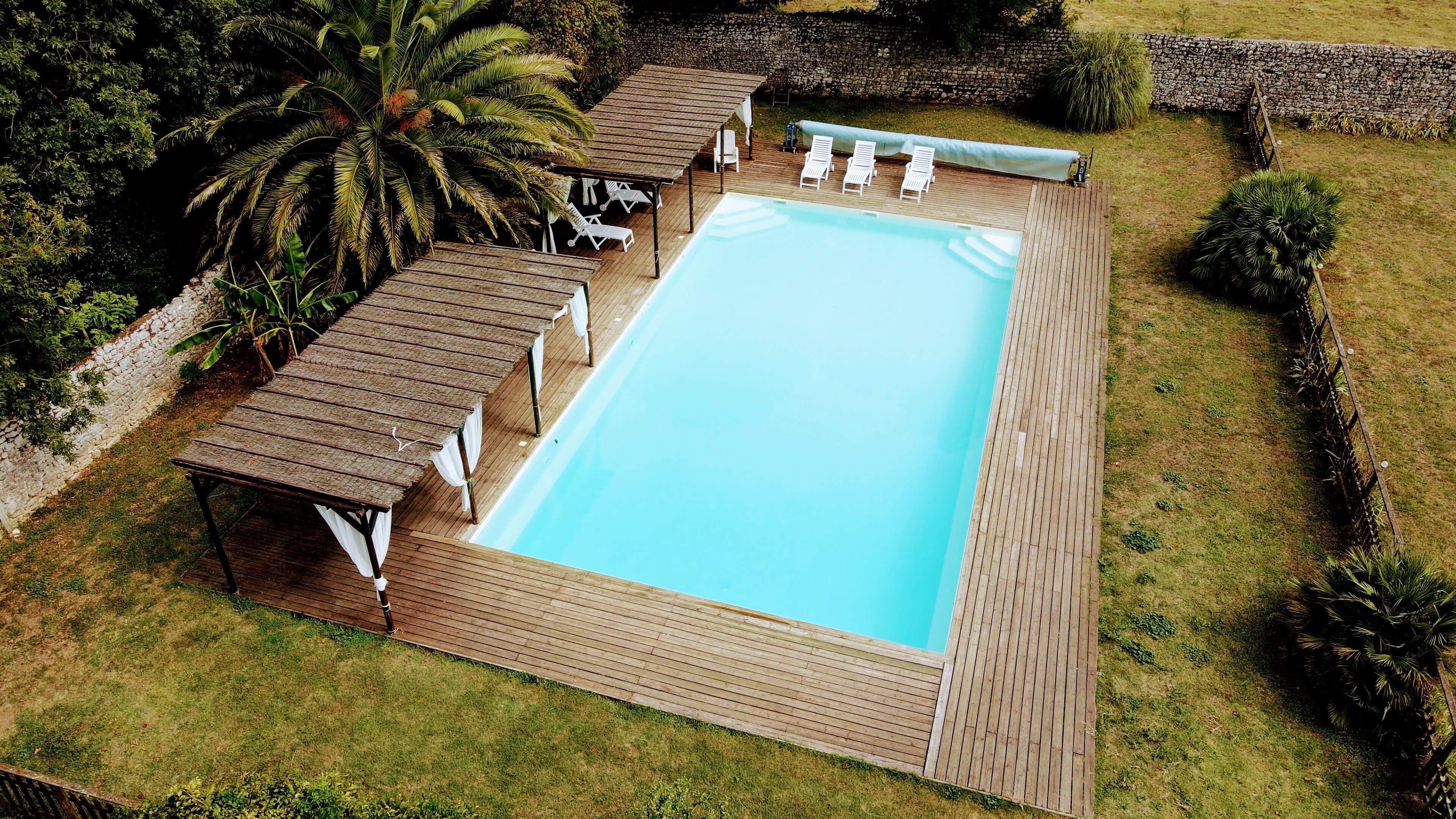 Ein rechteckiger Pool mit einer umlaufenden Holzterrasse, weißen Liegestühlen und einem überdachten Sitzbereich mit Sonnenschirmen.