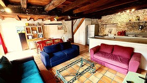Un salon rustique avec un canapé bleu et rose, un coin repas avec une nappe rouge et une cuisine dotée d’un mur en pierre et de poutres en bois.