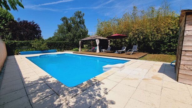 Ein rechteckiger Swimmingpool mit Fliesen umgeben von viel Grün und einer Holzterrasse mit Liegestühlen und einem Gazebo.