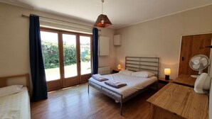 Ein Schlafzimmer mit Bett, Nachttischen, Schreibtisch, Lampe, Ventilator und Blick auf Grün durch das Fenster.