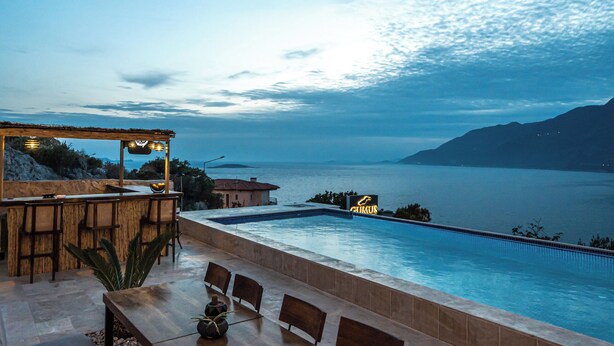 A poolside area with a wooden table and chairs, a bar, and a view of the sea. - GÜMÜŞ YAZAR  HOTEL (Kas)