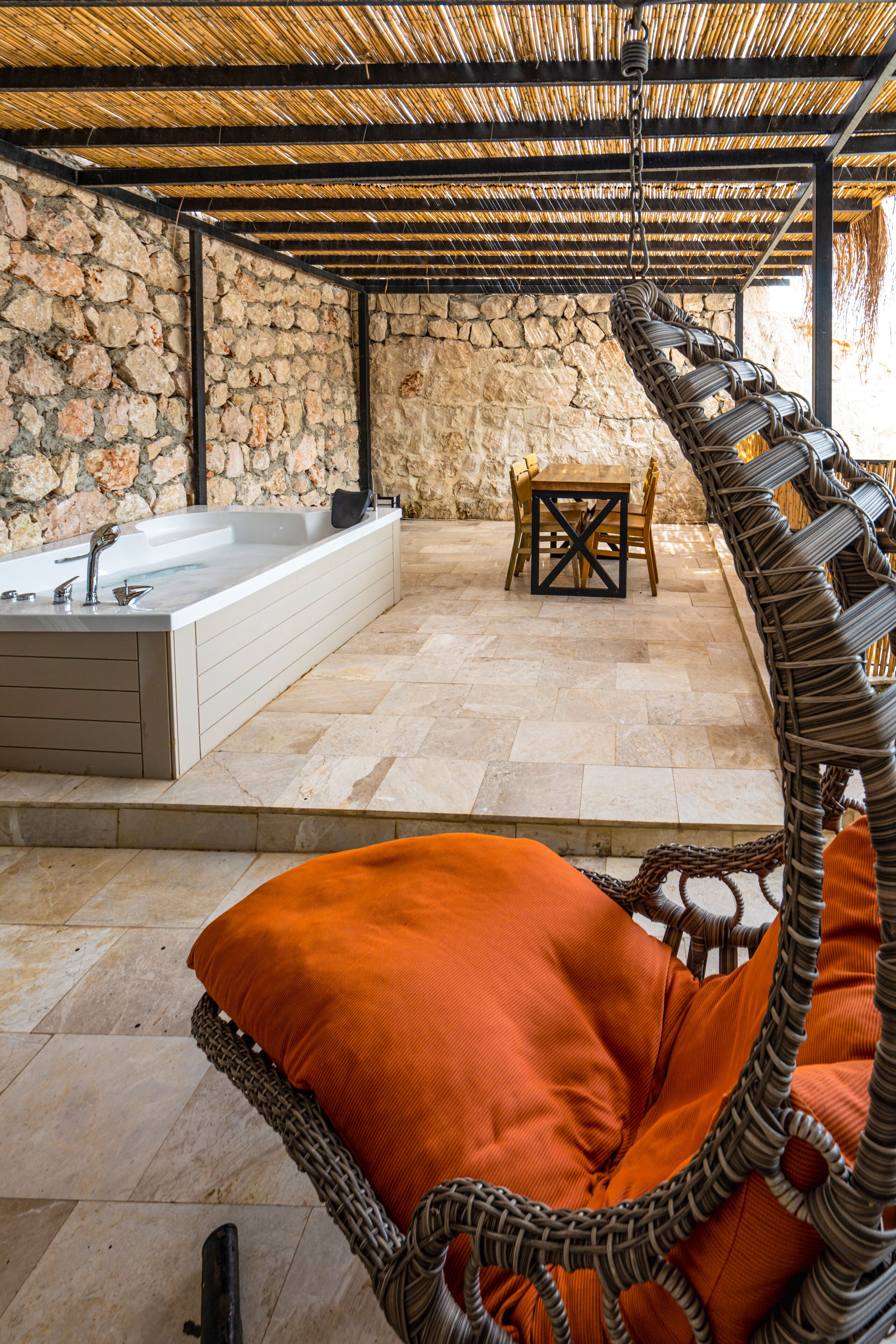 A stone-walled room with a bathtub, a wicker chair with an orange cushion, and a table with chairs.