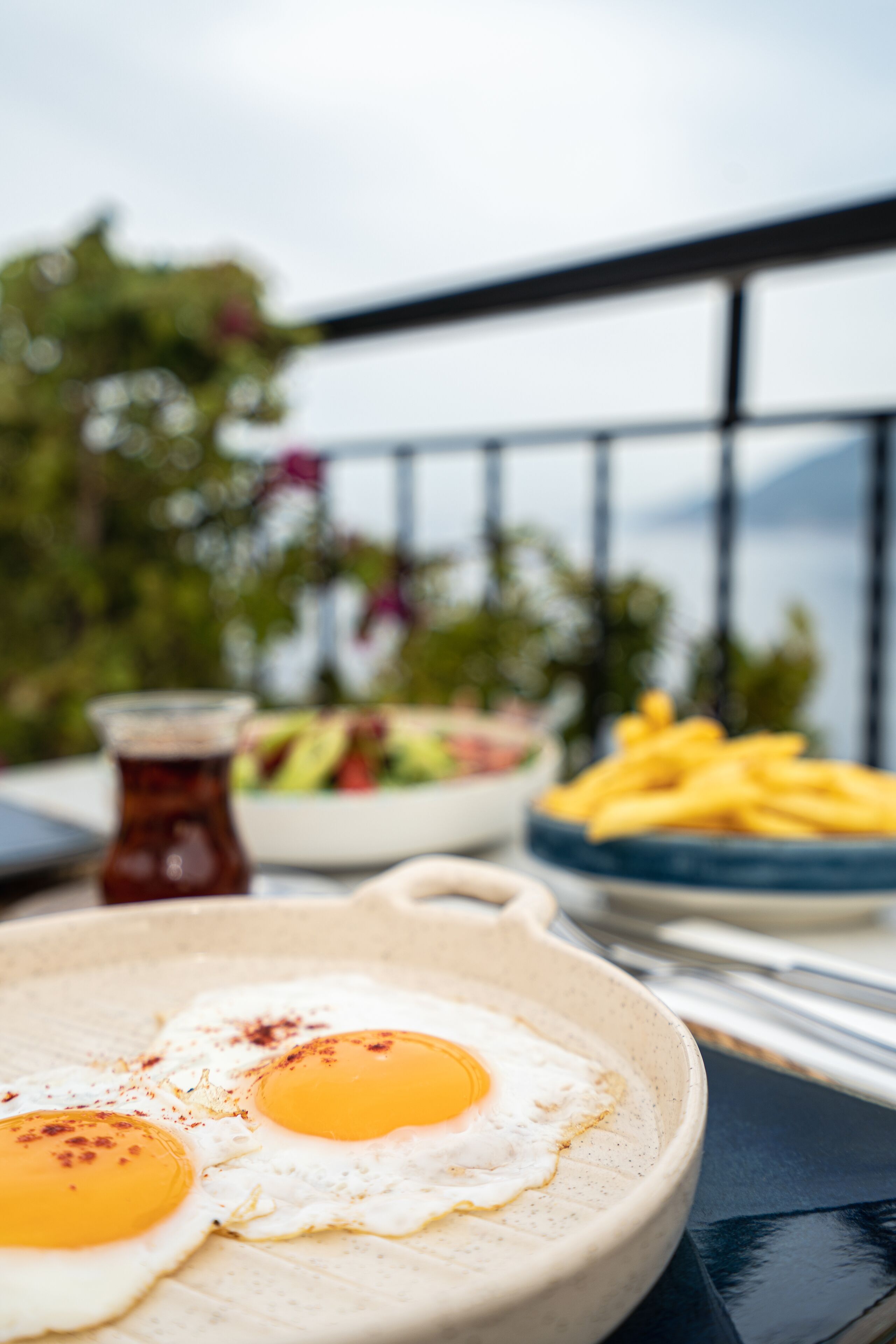 A breakfast setting with two sunny-side-up eggs, a bowl of fruit, and a plate of fries on a balcony with a view of greenery and a railing.