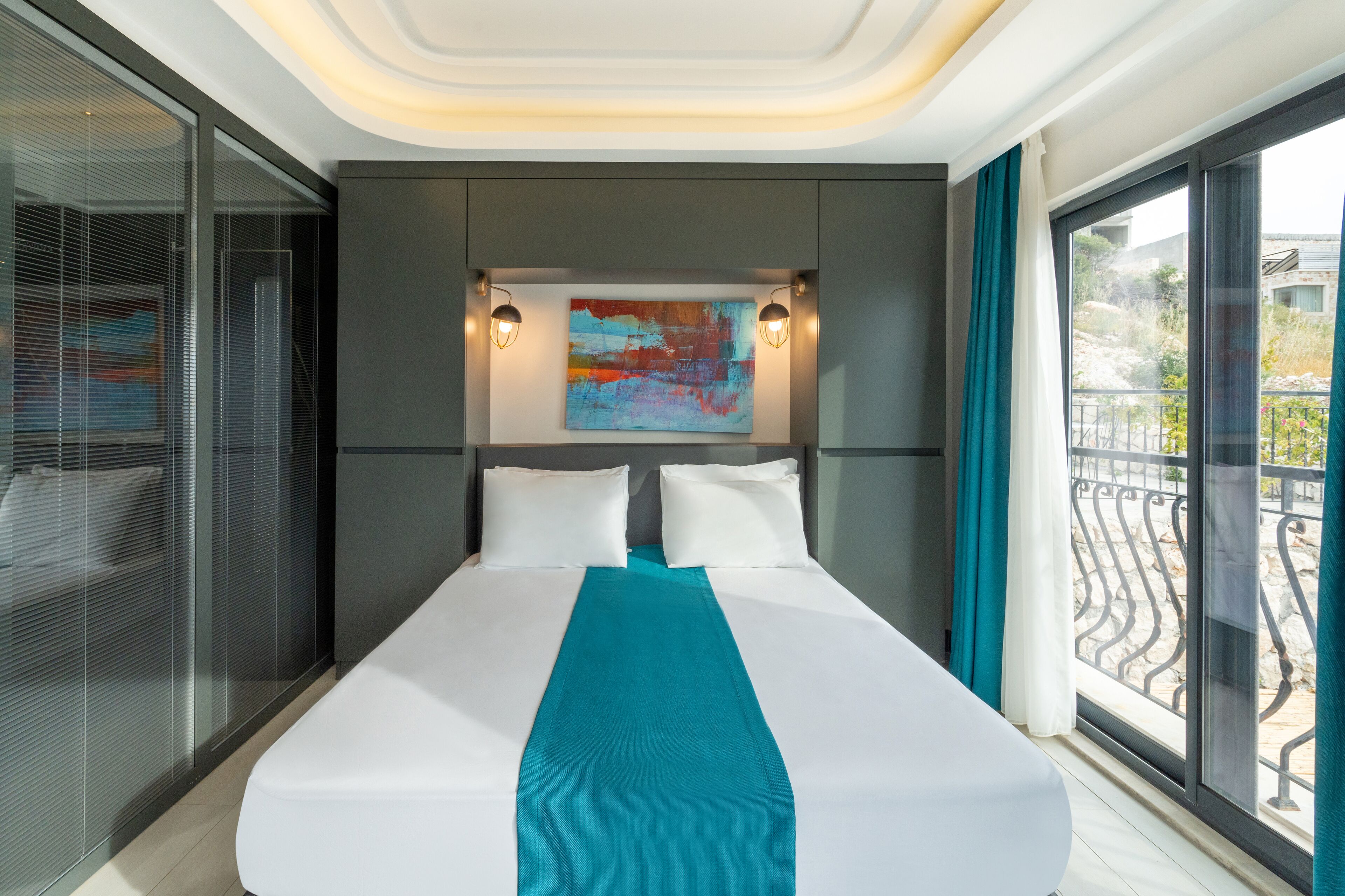 Una habitación de hotel moderna con una cama grande, un plaid color turquesa y un cuadro abstracto y colorido sobre la cama.