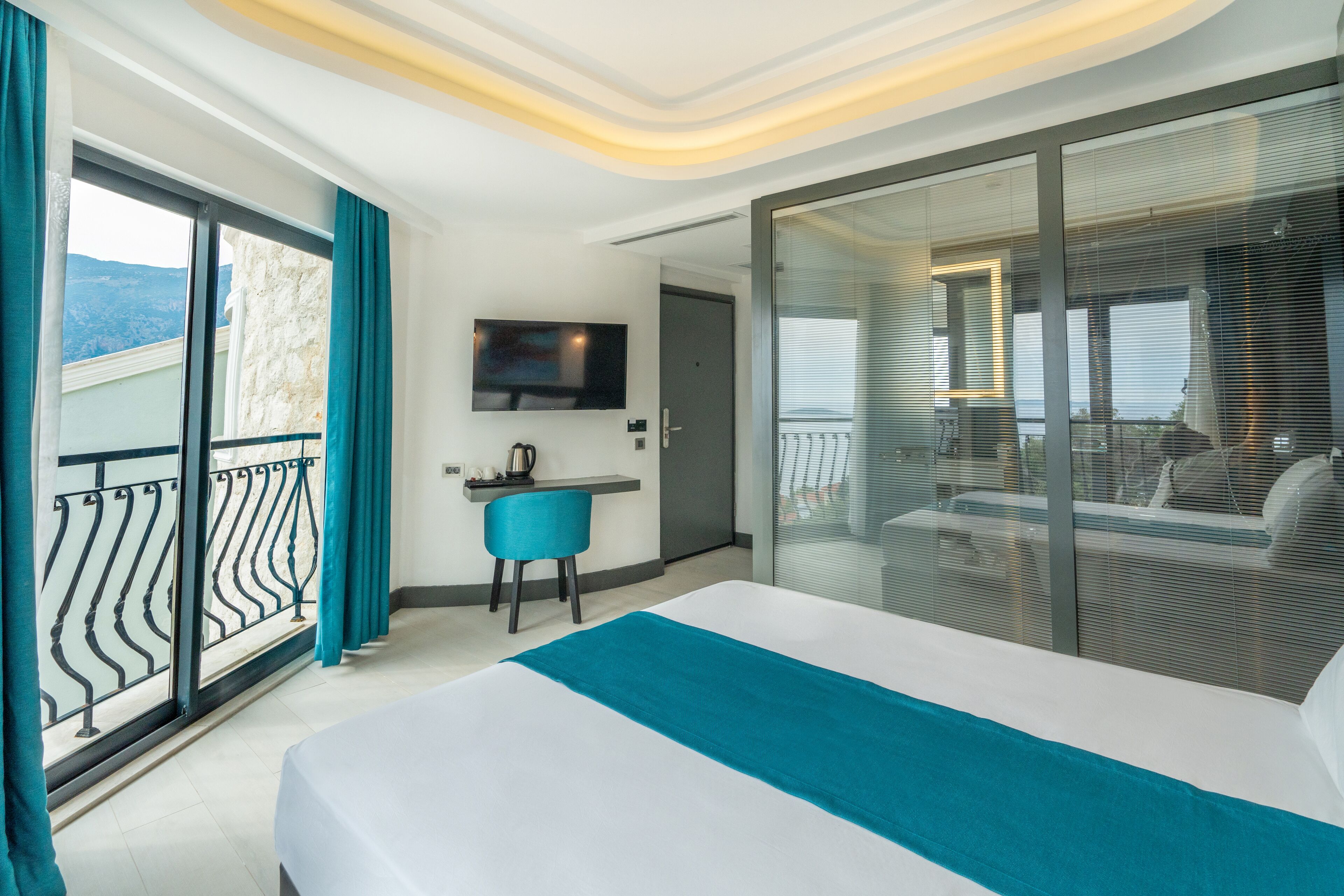 A modern hotel room with a large bed, a desk with a chair, a television, and a balcony with a view of the mountains.