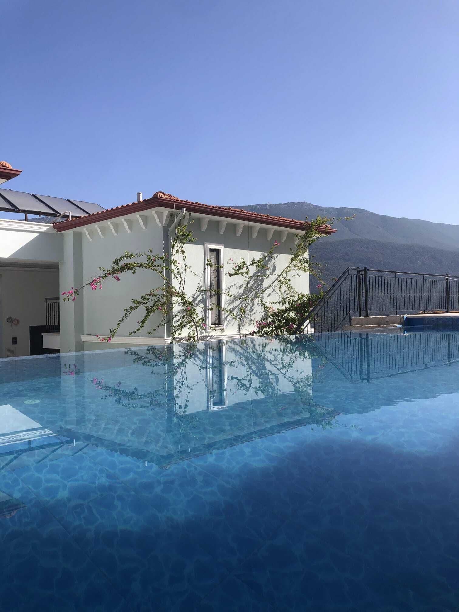 A clear swimming pool with a view of mountains in the background, a small building with a red-tiled roof, and a black metal fence surrounding the pool area.