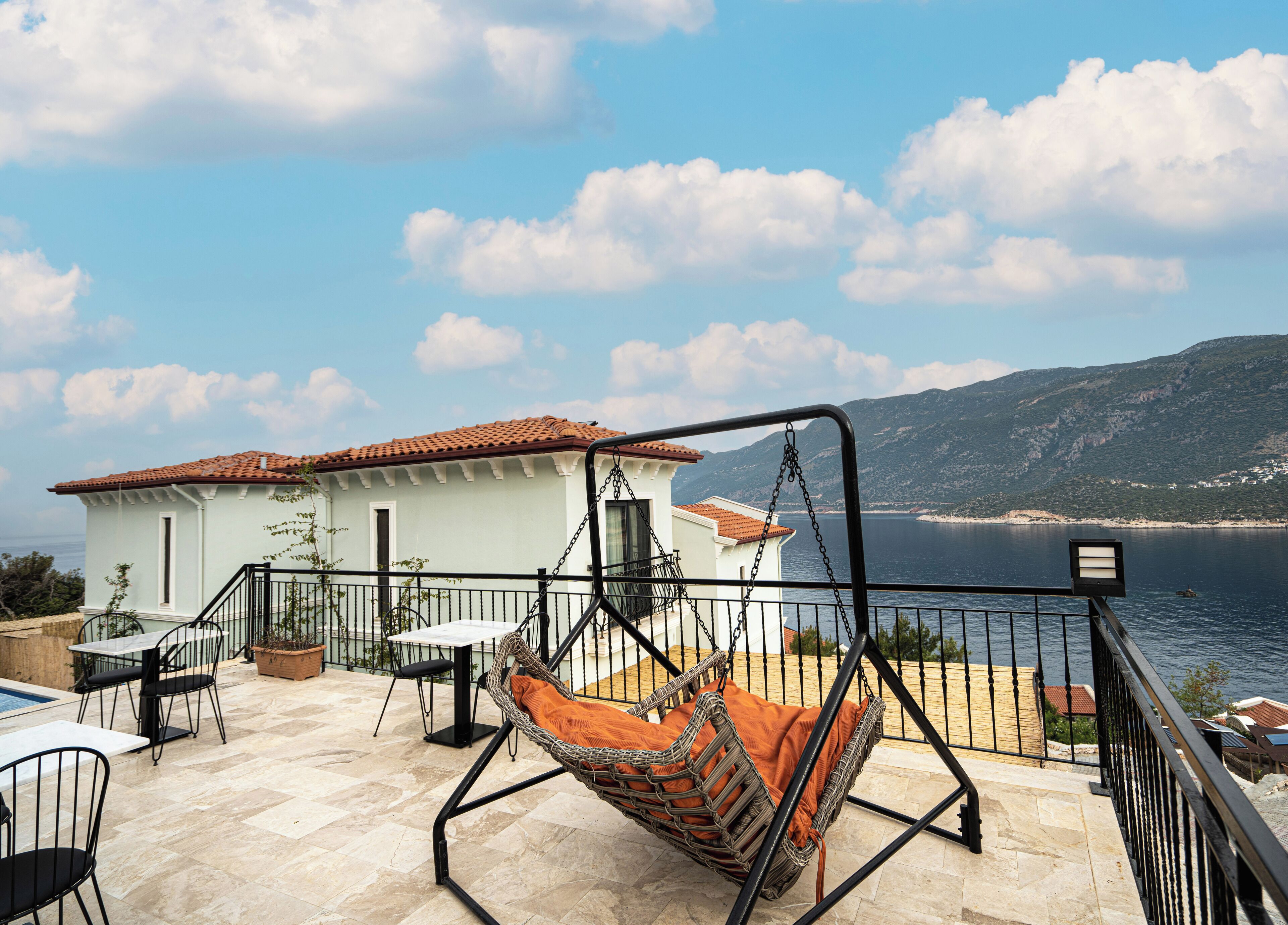 A patio with a swing chair, a table, and chairs, overlooking a body of water and a mountain.