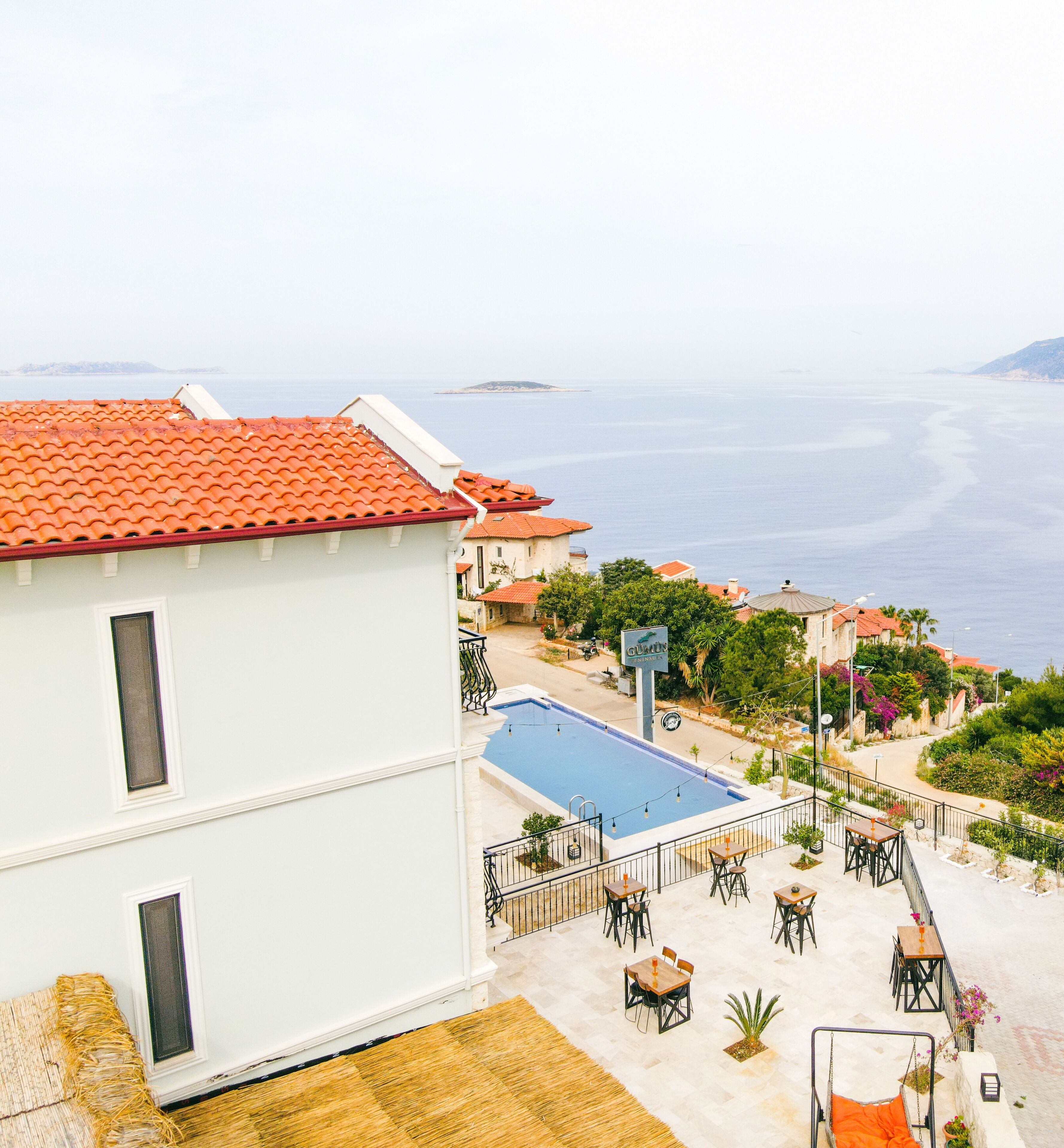 A white building with a red-tiled roof overlooking a swimming pool and outdoor seating area with tables and chairs.