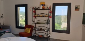 A room with a metal shelf unit displaying various kitchenware, a red chair with a yellow cushion, and a view of a green landscape through two windows. - House with a large pool and a magnificent view (La Londe-les-Maures)