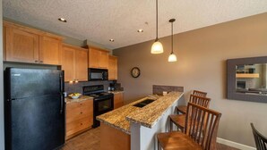 A kitchen with wooden cabinets, a black refrigerator, a microwave, a stove, and a granite countertop. There are two pendant lights and a clock on the wall. - 2 Bedroom Sooke Townhome (Sooke)