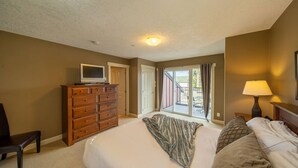 A bedroom with a large bed, a dresser, a TV on top, a chair, a lamp, and a view of the outside through a sliding door.